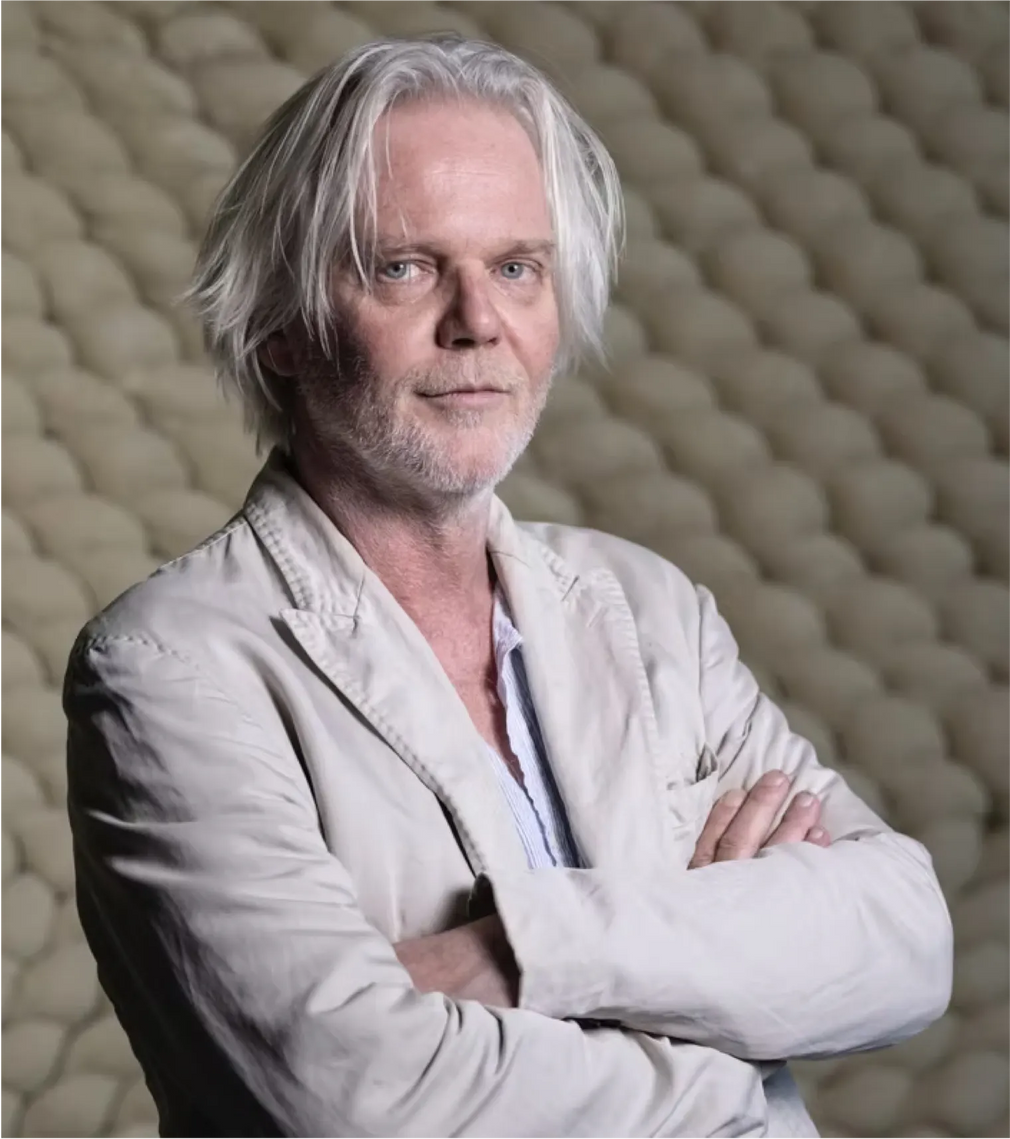 Front half-length portrait of Jürgen Strauss, founder and acoustic designer of STRAUSS ELEKTROAKUSTIK, looking directly ahead with crossed arms against a distinctive textured beige acoustic diffusion wall panel background.