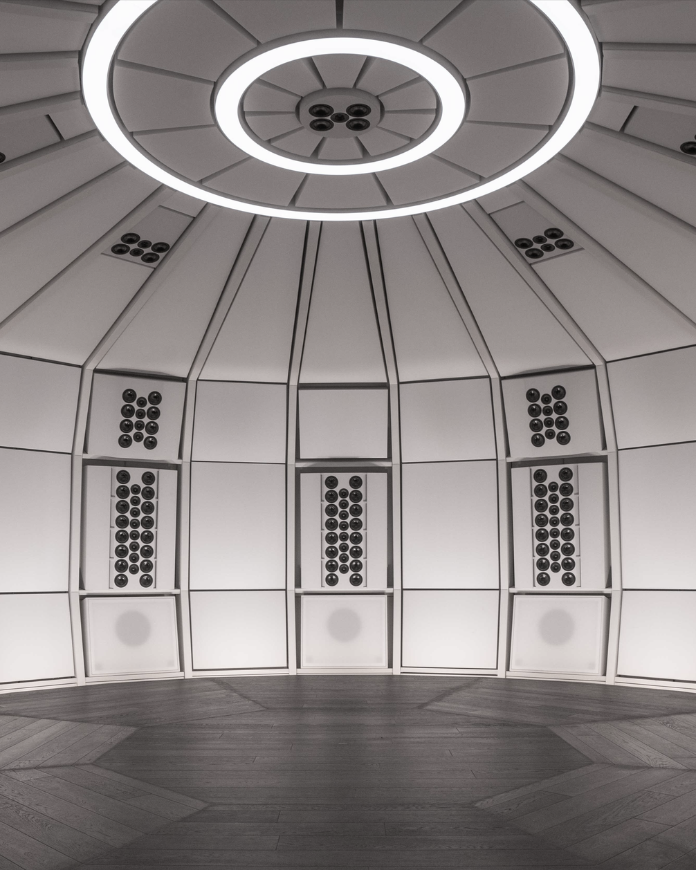 Vertical interior architectural view of the empty precision acoustic dome environment within THE LAB at SE MUSICLAB in Bern, Switzerland, showcasing the decoupled white geometric wall panels, dark wood flooring, and a distinctive multi-ring circular ceiling lighting fixture designed by Jürgen Strauss.