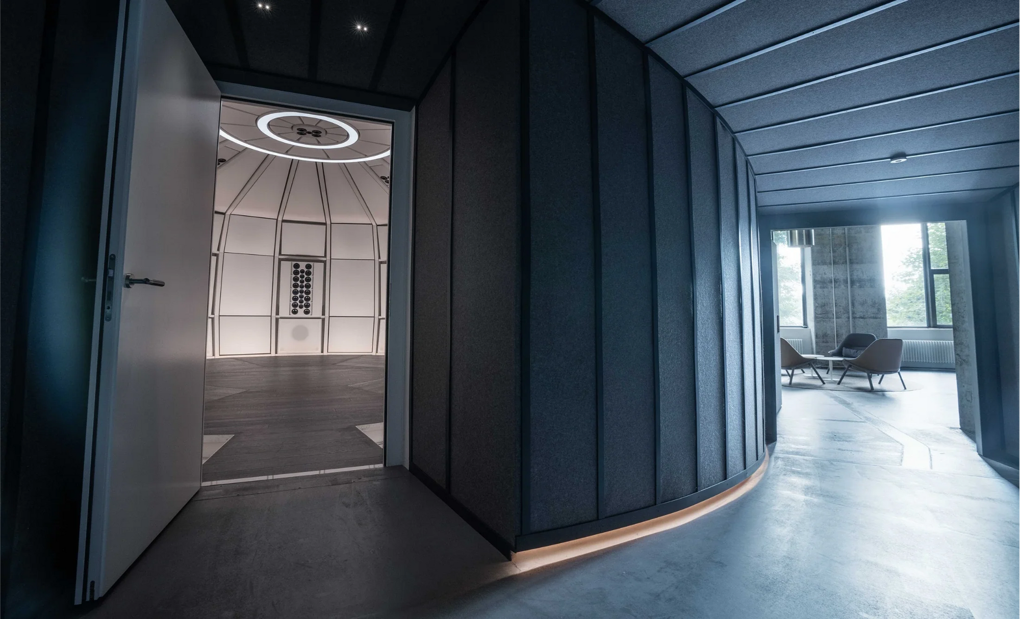 Interior architectural view from the hallway entry at SE MUSICLAB, showing the open doorway to the precision-engineered geometric dome of THE LAB listening room on the left, contrasting with a modern, sunlit lounge space and large windows on the right, highlighting the clean lines and advanced acoustic wall design.