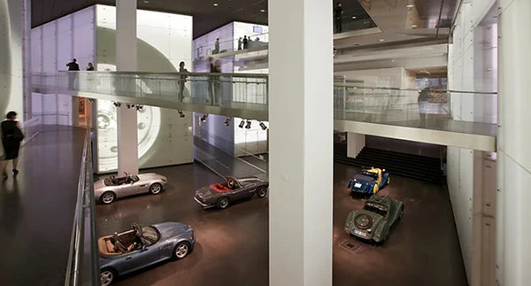 Wide high-angle interior view of the expansive central exhibition space at the BMW Museum in Munich, Germany, showcasing classic and modern sports cars on display across multi-level architectural platforms and elevated pedestrian walkways.