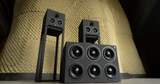 Front angle view of the STRAUSS ELEKTROAKUSTIK AP Series monitoring systems, showcasing the SE AP-1 on its custom floor stand, the SE AP-2, and the SE AP-SW Active-Passive reference subwoofer grouped together against a textured clay acoustic wall at the SE MUSICLAB.