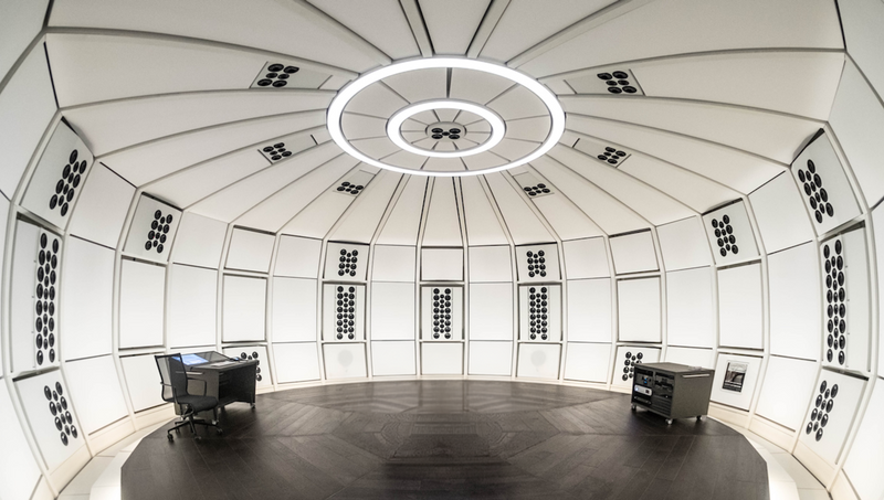 Wide interior view inside the precision acoustic dome environment of THE LAB at SE MUSICLAB in Bern, Switzerland, showing a central engineer's desk with a computer monitor and equipment racks positioned on dark wood flooring under a distinctive multi-ring circular ceiling light fixture.