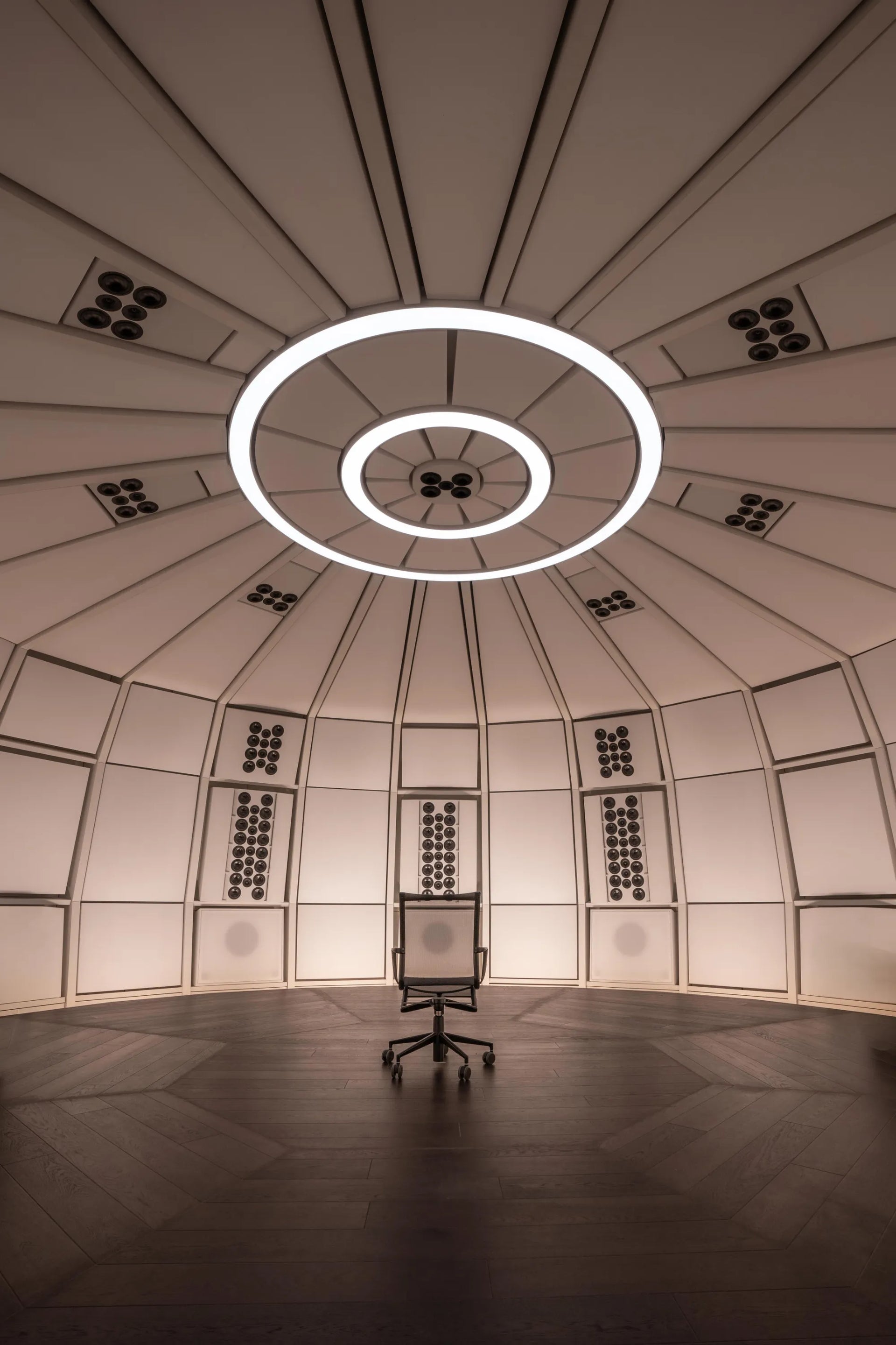 Vertical interior architectural view looking into the empty, precision-engineered geometric acoustic dome of THE LAB at SE MUSICLAB in Bern, Switzerland, highlighting a central listening chair set against a custom white decoupled modular wall system beneath a signature multi-ring circular ceiling lighting fixture.
