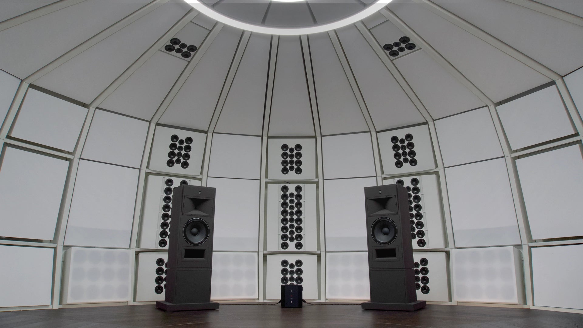 Interior architectural view of the precision-engineered acoustic environment of THE LAB at SE MUSICLAB in Bern, Switzerland, showcasing a pair of massive, custom floor-standing STRAUSS ELEKTROAKUSTIK MF-2.1 main monitors positioned against a custom white decoupled modular wall system beneath a signature multi-ring circular ceiling lighting fixture.