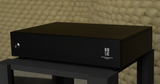 Front three-quarter perspective view of the STRAUSS ELEKTROAKUSTIK SE REQ-1S Reference Monitoring and Room Equalizer resting on a studio stand, showcasing the minimal dark chassis design, custom physical front panel layout with power indicator, and clean mechanical hardware alignment.